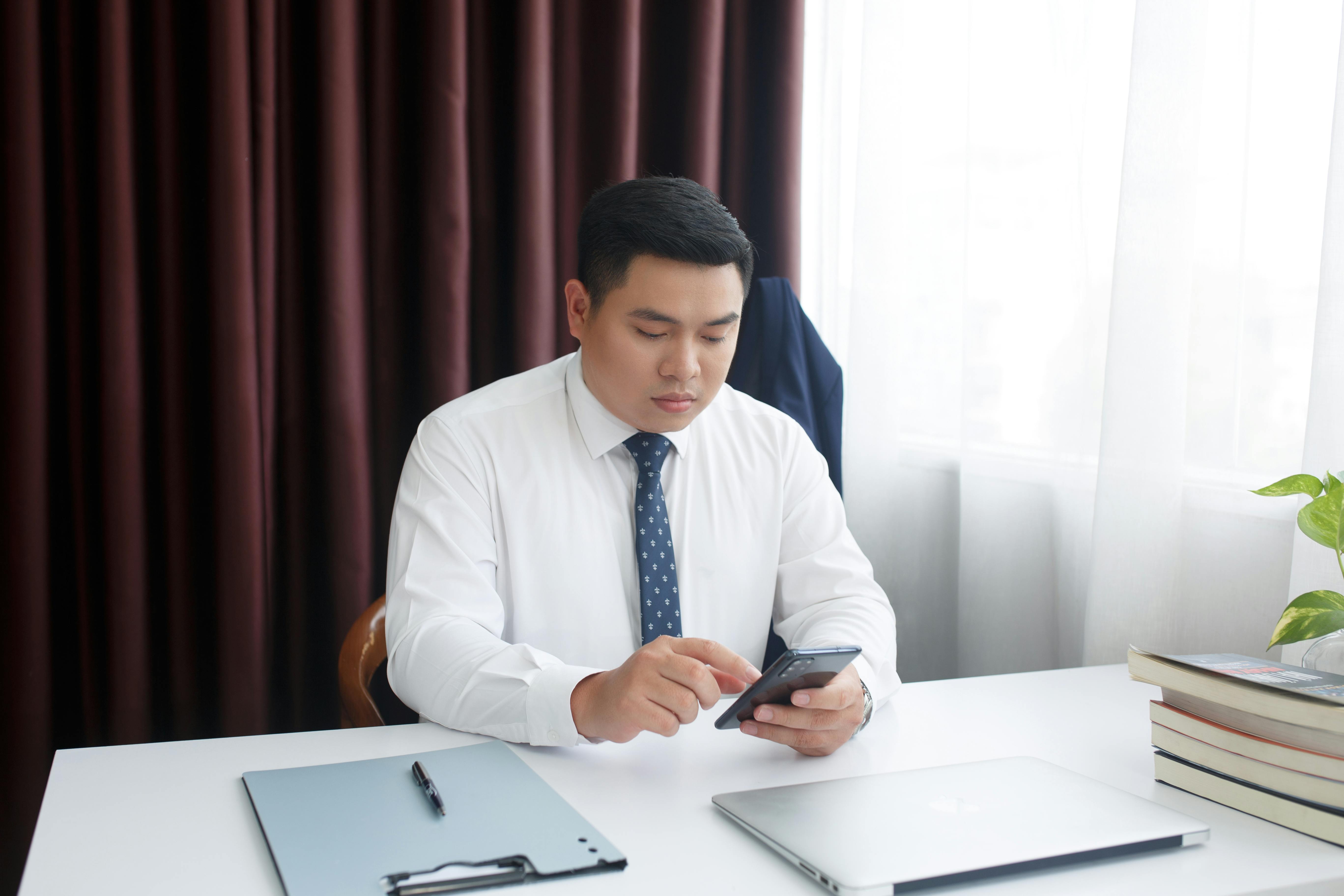 A businessman in a white shirt and tie using a smartphone at an office desk.